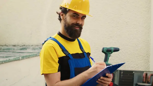 stock image Portrait of certified mechanic commissioned for outdoor air conditioner routine checkup, writing report on clipboard. Cheerful engineer doing hvac system inspection, looking for refrigerant leaks