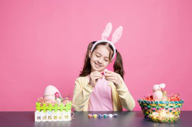 Tavşan kulaklı mutlu çocuk, Paskalya Pazar süsleri için yumurta boyuyor, geleneksel olarak el yapımı boyama süslemeleri yapıyor. Suluboya boyayla dekore eden neşeli kız..