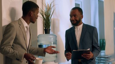 Cheerful african american businessmen enjoying nice water tank conversation during nightshift break. Joyful worker having cup of coffee relaxing while comunicating with colleague in office clipart