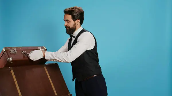 stock image Smiling bellboy carries trolley bags in studio, helping guests with luggage. Professional doorman illustrating role as hotel concierge staff in hospitality industry, tourism concept.