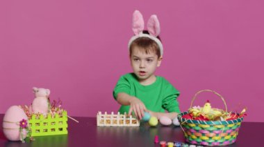 Tavşan kulaklı mutlu çocuk içi boyanmış yumurtalarla dolu sepeti düzenliyor, Paskalya Pazar kutlaması için şenlikli düzenlemeler yapıyor. Neşeli küçük çocuk sanattan ve sanattan hoşlanıyor. Kamera A.