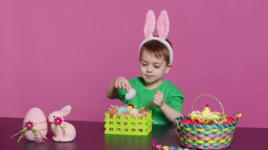 Heyecanlı küçük çocuk paskalya bayramı şenliğine hazırlanmak için sepetleri yumurta ve otla süsleyerek şenlikli düzenlemeler yapıyor. Tavşan kulaklı şirin bir bebek el işi yapıyor. Kamera A.