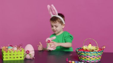 Doldurulmuş bir tavşan ve pembe bir yumurtayla oynayan sevimli küçük bir çocuk stüdyoda neşeli paskalya süsleri süslerken eğleniyor. Neşeli mutlu çocuk tatil hazırlıklarının keyfini çıkarıyor. Kamera A.