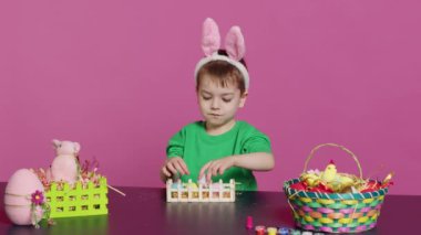 Mutlu çocuk paskalya düzenlemelerini el yapımı eşyalarla süslüyor, renkli yumurtaları sepete koyuyor. Neşeli küçük çocuk bahar bayramını kutluyor, pembe fon. Kamera A.