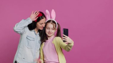Akıllı telefondaki fotoğraflara gülümseyen neşeli anne ve kız stüdyoda birbirlerini kucaklarken eğleniyorlar. Küçük çocuk ve annesi fotoğraflara poz veriyor, oynaşıyorlar. Kamera A.