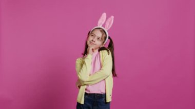 Paskalya bayramı için hediye ve yemek hayal eden gülümseyen küçük kız bahar tatili için heyecanlanıyor. Pembe arka planda poz veren tavşan kulaklı genç bir çocuk. Kamera B.