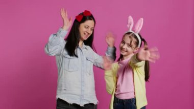 Gülümseyen anne ve küçük kız kameranın önünde el sallıyor, eğleniyor ve Paskalya bayramının tadını çıkarıyor. Stüdyoda annesiyle poz veren neşeli liseli kız merhaba diyor. Kamera B.