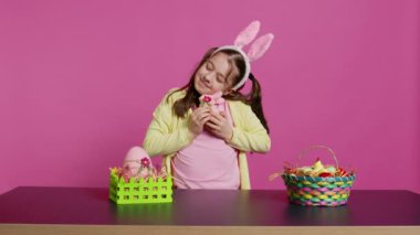 Neşeli gülümseyen kız oyuncak tavşan ve yumurtasına sarılıyor, el yapımı boyalı paskalya süslemeleriyle gurur duyuyor. Tavşan kulaklı, örgülü, şen şakrak bir çocuk. Kamera B.