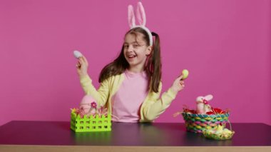 Hevesli genç bir kız, pembe arka planda boyalı paskalya yumurtalarını kullanarak kameranın önünde dikizcilik yapıyor. Neşeli, sevimli çocuk bahar bayramı şenliği için heyecanlanıyor. Kamera B.
