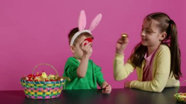 Stüdyoda paskalya geleneği için yumurta tokuşturan tatlı çocuklar, pembe arka planda sezonluk tatil oyunu oynuyorlar. Sevimli sevimli çocuklar, şenlikli dekorasyonlarla eğleniyorlar. Kamera B.