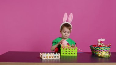 Paskalya Pazar kutlamaları için sepetler ve düzenlemeler hazırlayan neşeli gülümseyen çocuk, festival dekorasyonları için plastik çimenler ve sahte çiçekler koyuyor. Tavşan kulaklı mutlu bebek. Kamera B.