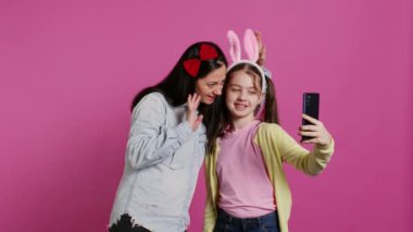 Akıllı telefondaki fotoğraflara gülümseyen neşeli anne ve kız stüdyoda birbirlerini kucaklarken eğleniyorlar. Küçük çocuk ve annesi fotoğraflara poz veriyor, oynaşıyorlar. Kamera B.