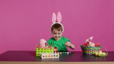 Sevimli küçük delikanlı Paskalya Pazar 'ı için el yapımı süsler yapıyor ve pul ve fırçalarla yumurta boyuyor. Sanat malzemelerini boyamak ve dekore etmek için kullanan mutlu genç bir çocuk. Kamera B.