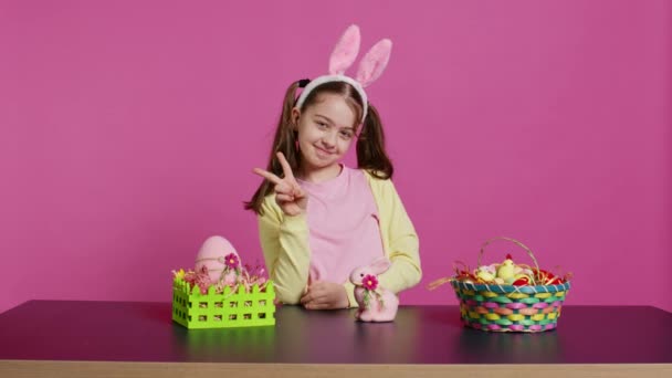 在演播室里表现出和平的快乐纯真的孩子 戴着小兔子耳朵 带着节日装饰品在桌子上庆祝复活节 精力充沛 有创意的快乐女孩 摄像头B — 图库视频影像