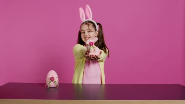 用兔子耳朵微笑幼儿 展示她手工制作的复活节装饰品 展示一个手工制作的彩蛋和兔子 年轻甜美的女学生拿着节日装饰品 摄像头B — 图库视频影像