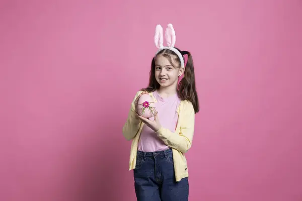 可爱的小女孩 长着小兔子的耳朵 在镜头前拿着一个粉红的彩蛋 在彩色的背景上展示她手工制作的东方装饰 年轻可爱的女孩展示传统的装饰品 — 图库照片
