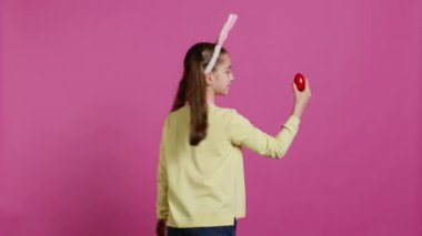 Coşkulu küçük bir çocuk, Paskalya bayramı için kırmızı boyalı yumurtasını sunarak kameranın önünde dönüyor. Tavşan kulaklı neşeli bir çocuk. Dönüyor ve piruet yapıyor. Kamera B.