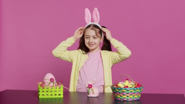 彼女の頭の上にバニーの耳を置いて ピンクの背景にこんにちは イースターの日曜日の休日のイベントのための装飾を作成するかわいい喜びの子供 カメラ — ストック動画
