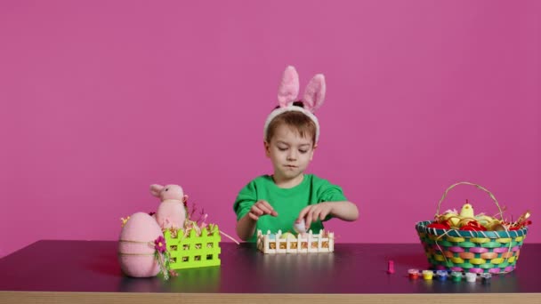 幸せな子供は 手作りのアイテムでイースターのアレンジを飾り バスケットにカラフルな卵を配置しました 春のお祭りを祝う楽しい少年 ピンクの背景 カメラ — ストック動画