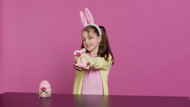 用兔子耳朵微笑幼儿 展示她手工制作的复活节装饰品 展示一个手工制作的彩蛋和兔子 年轻甜美的女学生拿着节日装饰品 相机A — 图库视频影像