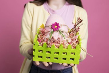 Küçük neşeli çocuk sepetinde el yapımı paskalya süslemeleri gösteriyor pembe arka planda renkli yumurtalar sunuyor. Stüdyoya özgüvenle poz veren pozitif küçük kız, tavşan kulaklı. Kapat..