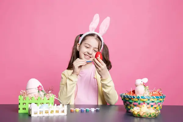 カラフルなデザインを描いた小さな熱狂的な子供は イースターのお祭り ピンクの背景に備えるために水彩で卵を描きます 彼女がテーブルでアートを作成するようにリラックスする若い女の子 — ストック写真