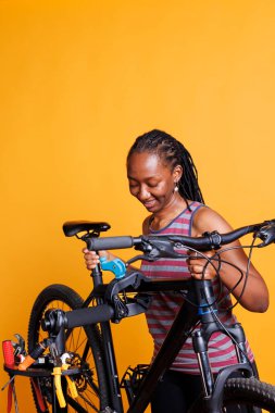 Active healthy female securing bike on repair-stand for repairing against yellow background. Image showing sporty black woman adjusting bicycle height, ready for yearly maintenance. clipart