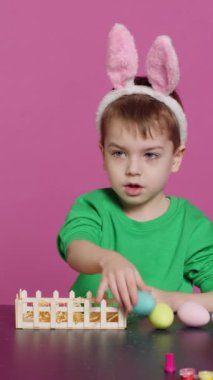 Tavşan kulaklı dikey videolu çocuk içi boyanmış yumurtalarla dolu sepeti düzenliyor ve Paskalya Pazar kutlamaları için şenlikli düzenlemeler yapıyor. Neşeli küçük çocuk sanattan ve sanattan hoşlanıyor.