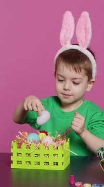 Dikey Video, Paskalya bayramı şenliğine hazırlanmak için sepetleri yumurta ve otla süsleyerek şenlikli düzenlemeler yapan küçük çocuğu heyecanlandırdı. Tavşan kulaklı şirin bir çocuk iş yapıyor.