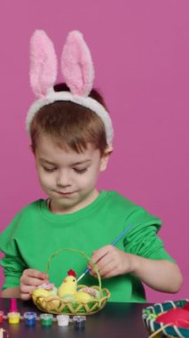 Dikey Video Mutlu küçük çocuk Paskalya Pazar kutlamalarına hazırlanmak için sepette yumurta ve civciv düzenliyor, stüdyoda şenlikli dekorasyonlar yaratıyor. Küçük çocuk boyama yaparken eğleniyor. Kamera A.