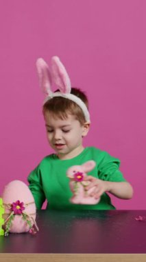 Doldurulmuş bir tavşan ve pembe bir yumurtayla oynayan sevimli küçük bir çocuk stüdyoda neşeli paskalya süsleri süslerken eğleniyor. Neşeli mutlu çocuk tatil hazırlıklarının keyfini çıkarıyor. Kamera B.