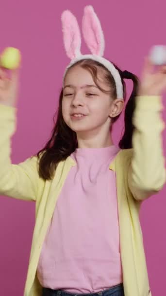スタジオでブーケゲームをプレイする垂直ビデオアドラブルな少女は ピンクの背景に対して彼女の手作りの着色されたイースターエッグを示しています バニー イヤーが バカにしてる 陽気な遊び心の子供 カメラ — ストック動画