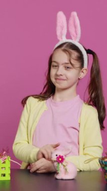 Vertical Video Cheery 'nin masum çocuğu stüdyoda barış işareti gösteriyor ve tavşan kulağı takıyor, Paskalya bayramını bir masada şenlikli süslerle kutluyor. Yaratıcılığı olan mutlu enerjik kız.