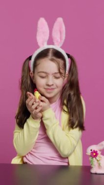 Dikey Video Neşeli küçük kız stüdyoda neşeli paskalya süslemeleriyle oynuyor, bir piliç, tavşan ve yumurta ile düzenlemeler yapıyor. Gülümseyen sevimli bebek tavşan kulaklarıyla renkli süsler gösteriyor.