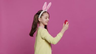 Coşkulu küçük bir çocuk, Paskalya bayramı için kırmızı boyalı yumurtasını sunarak kameranın önünde dönüyor. Tavşan kulaklı neşeli bir çocuk. Dönüyor ve piruet yapıyor. Kamera A.