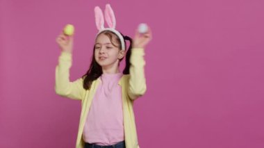 Stüdyoda el yapımı renkli paskalya yumurtalarını pembe arka planda gösteren küçük sevimli bir kız. Tavşan kulaklı neşeli, şakacı çocuk. Kamera A.