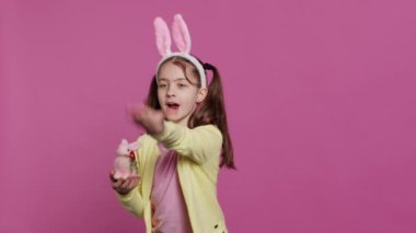 Tavşan kulaklı sevimli küçük kız stüdyoya hava öpücüğü gönderiyor, Paskalya tatili için doldurulmuş pembe bir tavşan oyuncağı sunuyor. Gülümseyen enerjik ergen pozitif ve tasasız hissediyor. Kamera A.
