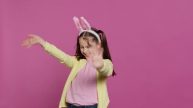 Sevimli şirin çocuk tavşan kulağı takıyor ve kameraya el sallıyor. Pembe arka planda Paskalya Pazar kutlamasının tadını çıkarıyor. Saçları örgülü, güler yüzlü liseli kız merhaba diyor. Kamera A.