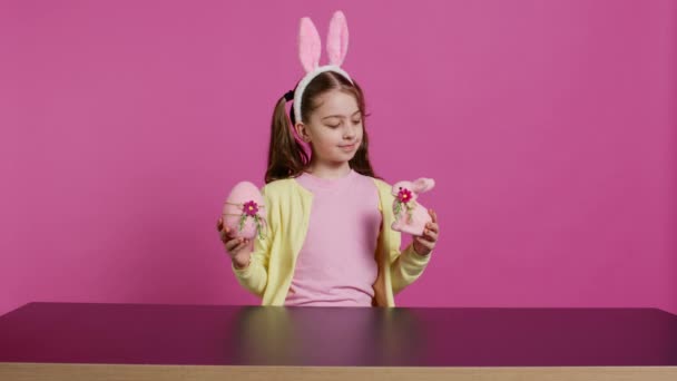 カメラにカラフルな装飾の装飾を施した小さな陽気な少女が手作りのお祝いのアレンジを作ります イースターの日曜日に興奮する若い子供は ウサギと卵を持っています カメラ — ストック動画