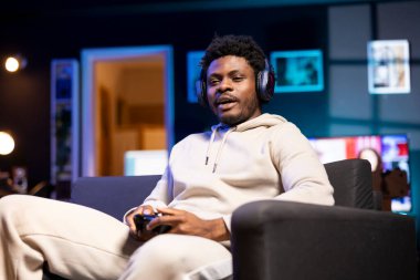 BIPOC oyuncusu evde dinleniyor, koltuğa oturuyor, oyun oynamak ve boş zamanların tadını çıkarmak için heyecanlı. Adam oturma odasında kanepede oturuyor, oyun konsolunda video oyunu oynuyor, işten sonra dinleniyor.