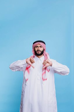 Geleneksel giysiler giyen hoşnutsuz Arap başparmakları aşağıda poz veriyor ve ciddi bir ifadeyle kameraya bakıyor. Müslüman adam Thombe ve Gutra giyinmiş ve onaylanmamış jest yapıyor.
