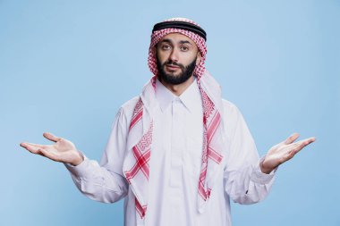 Müslüman gibi giyinmiş, başörtüsü takan, ayakta duran ve kameraya bakarken omuz silken bir adam. Arap biri kollarını açmış stüdyo portresiyle tereddüt ediyor.