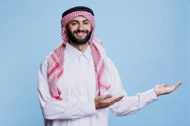 Geleneksel cüppeli Müslüman adam, stüdyo portresiyle sol tarafı işaret ediyor. Arap ürün reklamı ve neşeli bir ifadeyle kameraya bakıyor.