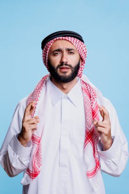 Geleneksel kıyafetler giymiş Müslüman bir adam parmaklarını çapraz yapıp umut dolu bir ifadeyle kameraya bakıyor. Araplar batıl inançlı portrelerle şans dilemek için dua ediyorlar.