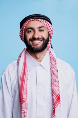 İslami beyaz tobe ve damalı ghutra başlıklı stüdyo portresi giymiş neşeli Müslüman adam. Mutlu Arap insan ayakta durup kameraya kaygısız bir ifadeyle bakıyor.