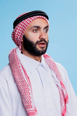 Geleneksel başörtüsü takmış Müslüman adam gözlerini kaçırıp ağzını açtı. Arap ghutra ve thobe giysileri giyen kişi şaşırmış yüz ifadesiyle poz veriyor.