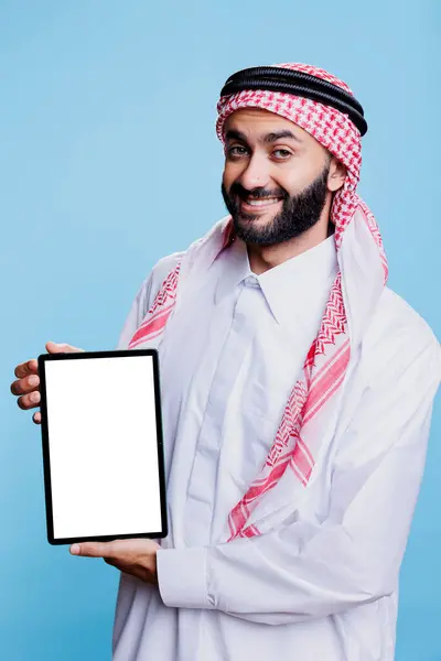 白い空白のスクリーンのモックアップとカメラを見ているデジタルタブレットを保持する伝統的な服装の笑顔のアラブ アプリケーション広告のためのポータブルガジェットの空のタッチスクリーンを促進するムスリム男性 — ストック写真