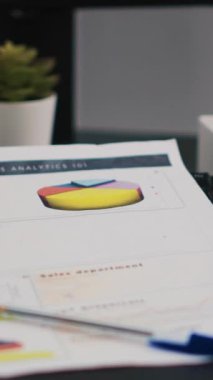 Masanın üzerinde, boş bir ofiste kağıt bardak kahvenin yanında duran dikey video finans raporu. Yakın çekim. Çalışma alanındaki sıcak içecek masasının yanında ekonomik grafikler ve pasta grafikleri içeren şirket dosyaları