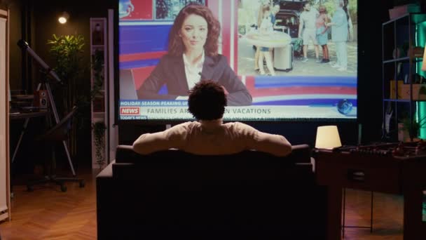 ホームシアターの男性は 家族旅行でニュース放送の報道を示す巨大なテレビ画面を見ています ワイドスクリーンテレビセットでストリーミングサービスを視聴するコードカッターで撮影したズーム — ストック動画