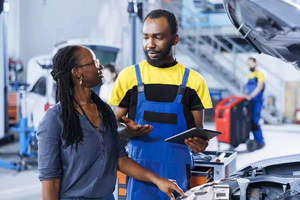 stock image Mechanic at auto repair shop conducts annual vehicle checkup, informing customer about needed motor oil replacement. BIPOC garage worker talks with customer after finishing car inspection